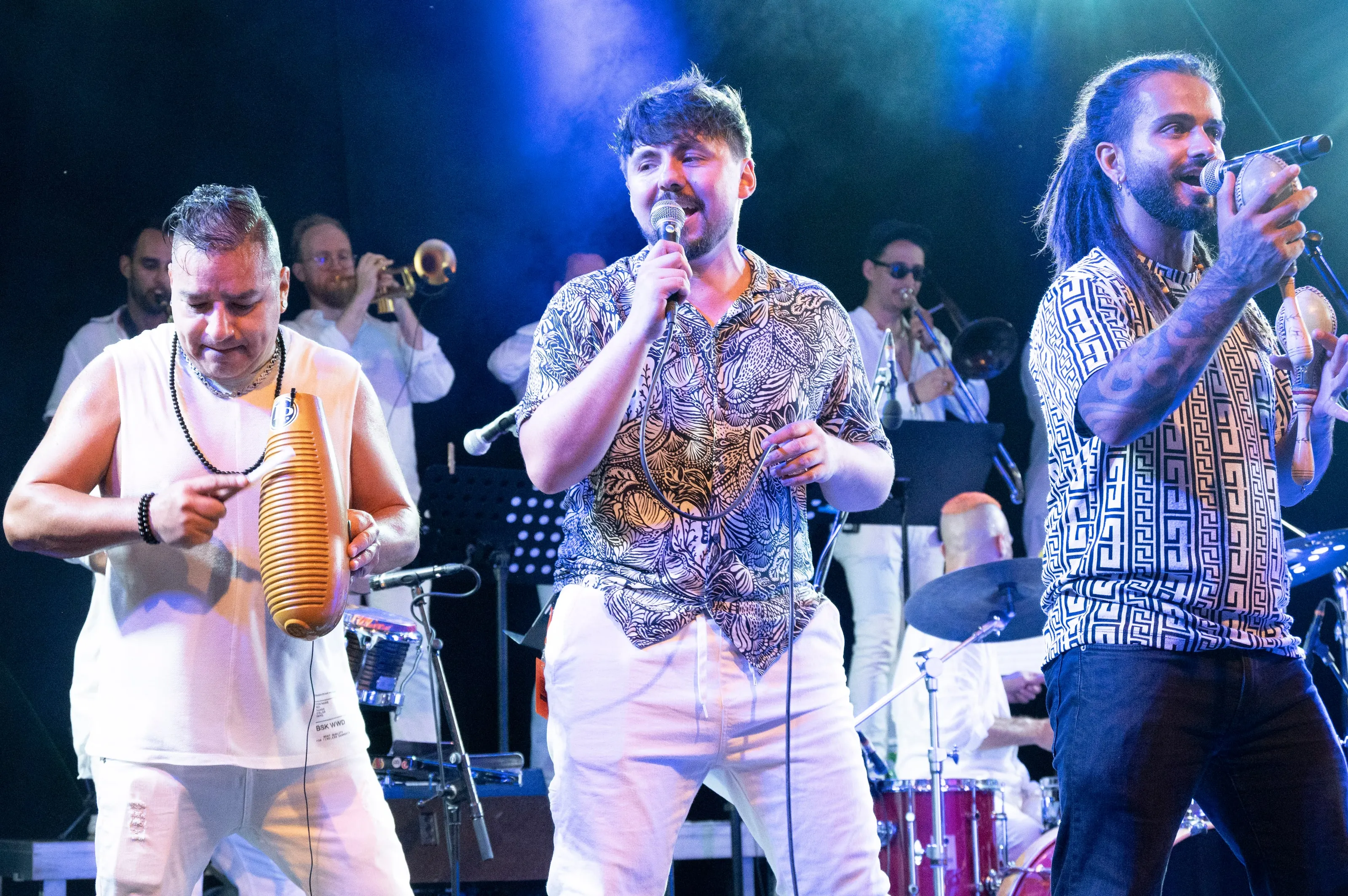 Güiro et maracas en main, trois chanteurs mènent le son dans une lumière bleue, cuivres et batterie en arrière-plan