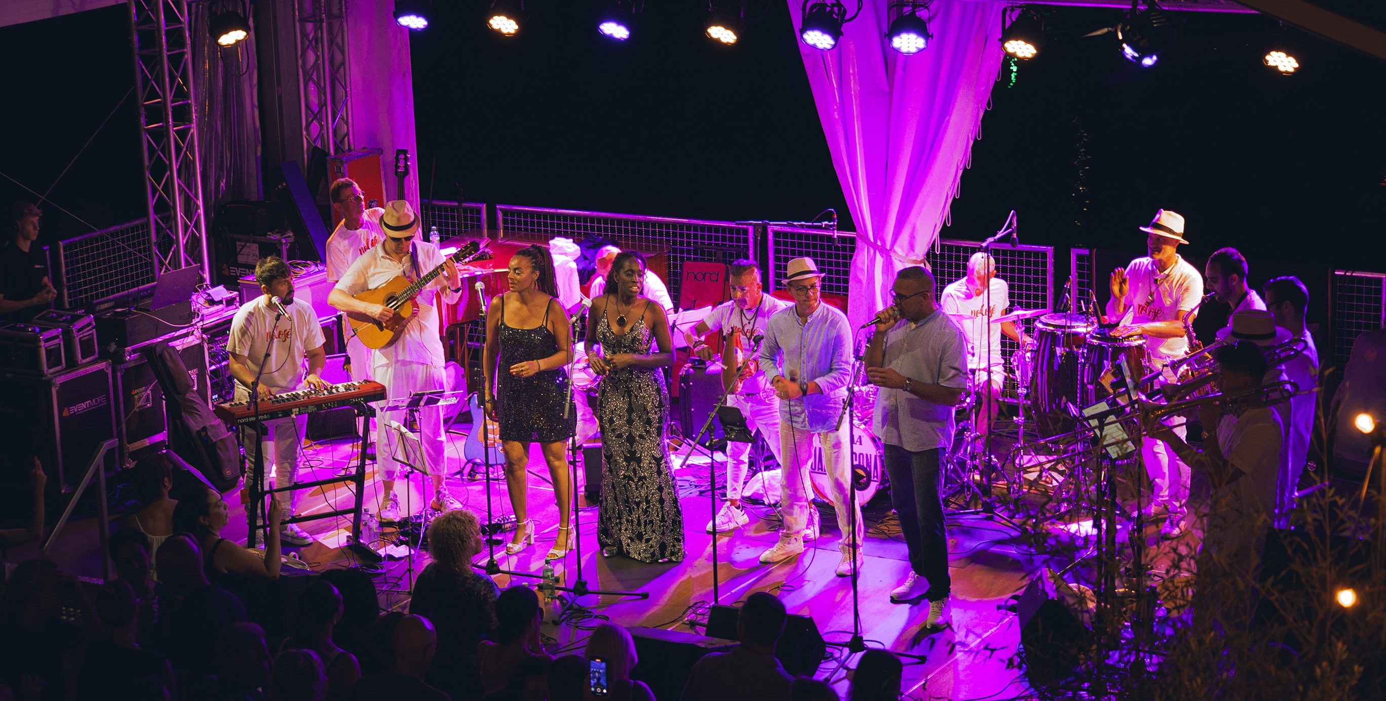 L'orchestre au complet en blanc sous les lumières violettes d'une scène couverte, le public attablé au premier rang